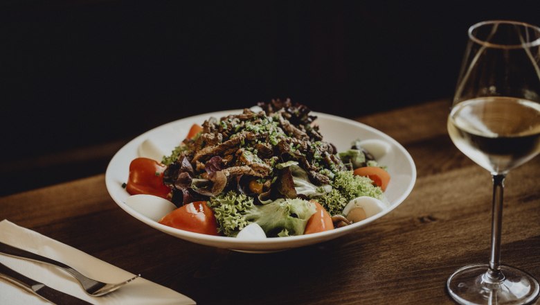 Heiligenkreuzer Spezialsalat, © Niederösterreich Werbung/Sophie Menegaldo Ein Teller mit gemischtem Salat, Tomaten, Eiern und Fleischstreifen auf einem Holztisch, daneben ein Glas Weißwein.