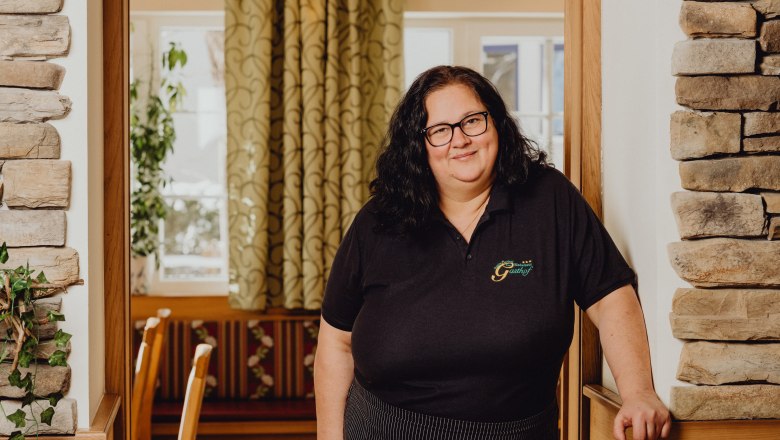 Wirtin Silke Perthold, © Niederösterreich Werbung/Klaus Engelmayer Eine Frau mit Brille steht in einem gemütlichen Gasthof mit Steinwänden und Vorhängen.