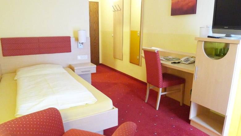 Roter Hahn, © Gasthof-Pension Böck Ein Hotelzimmer mit Einzelbett, rotem Teppich, Schreibtisch und Stuhl. An der Wand hängt ein Bild, daneben ein Fernseher auf einem Schrank.