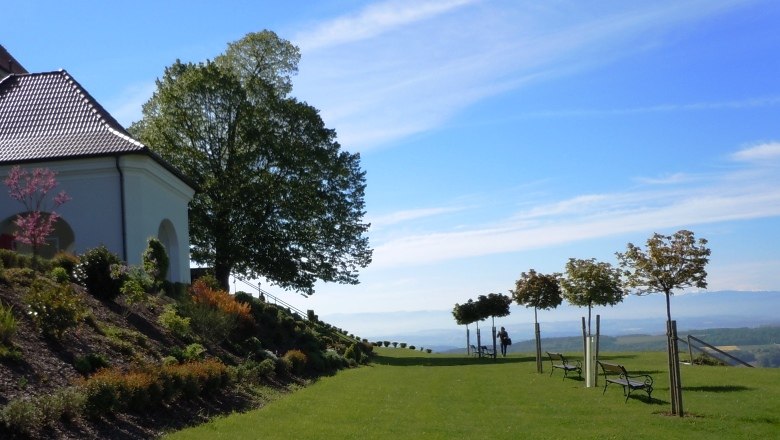 Klostergarten Maria Taferl, © "Natur im Garten" Schaugärten, Litschauer Ein gepflegter Garten mit Bäumen und Bänken, neben einem Gebäude mit rotem Dach, unter blauem Himmel.
