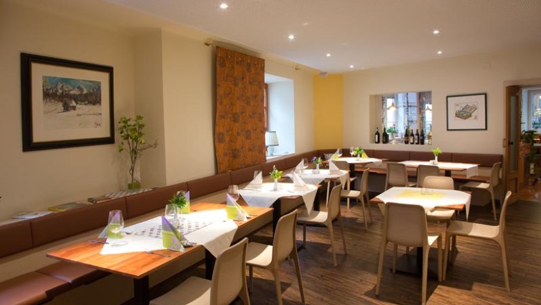 Speiseraum, © Gerhard Böhm Ein moderner Speiseraum mit gedeckten Tischen, Stühlen und Dekorationen. An den Wänden hängen Bilder, und es gibt eine Fensterbank mit Weinflaschen.