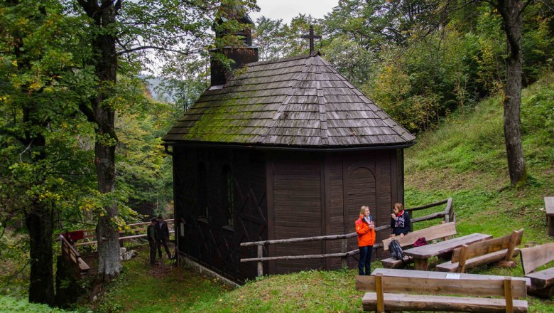 Hölzerne Kirche, © zur Verfügung gestellt: Gemeinde Schwarzenbach an der Pielach Eine kleine, hölzerne Kirche im Wald mit zwei Personen und Holzbänken im Vordergrund.