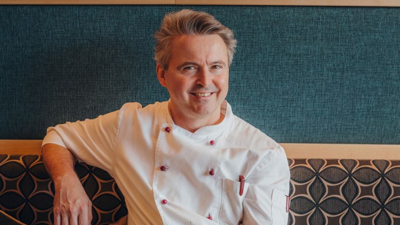 Wirt Hermann Froschauer, © Niederösterreich Werbung/Daniela Führer Ein lächelnder Mann in weißer Kochjacke sitzt auf einer gepolsterten Bank vor einer gemusterten Wand.