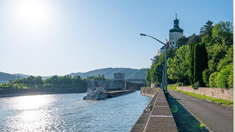 An der Donaulände, © Guestia GmbH Fluss mit Schiff und Gebäude am Ufer, sonniger Himmel.
