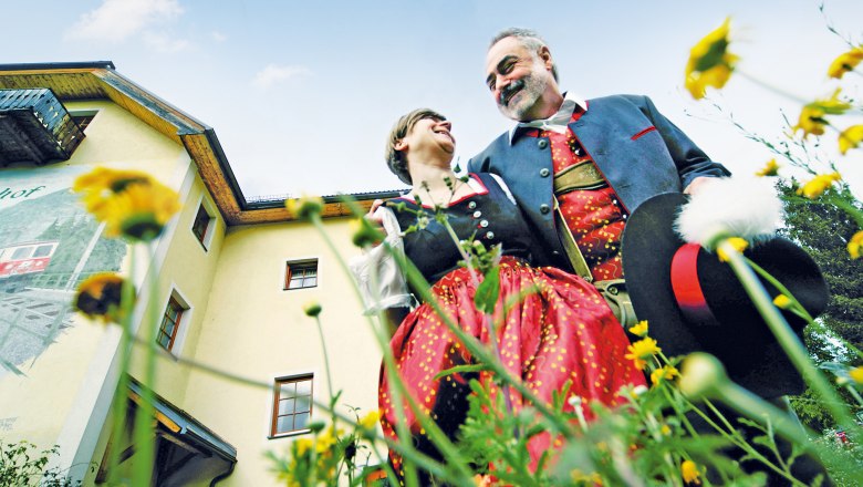 Naturhotel Steinschalerhof, © weinfranz.at Ein Paar in traditioneller Tracht steht vor einem gelben Gebäude, umgeben von Blumen, und lächelt sich an.