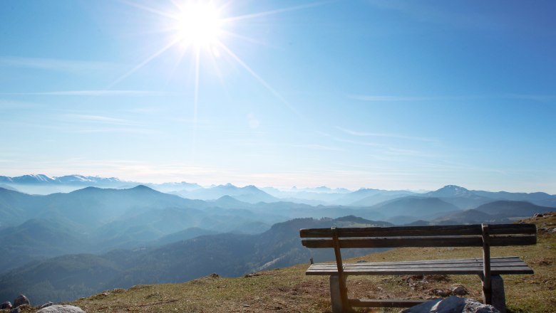 Gemeindealpe Mitterbach, © weinfranz.at Holzbank auf einem Berg mit Blick auf eine Berglandschaft und die Sonne am Himmel.
