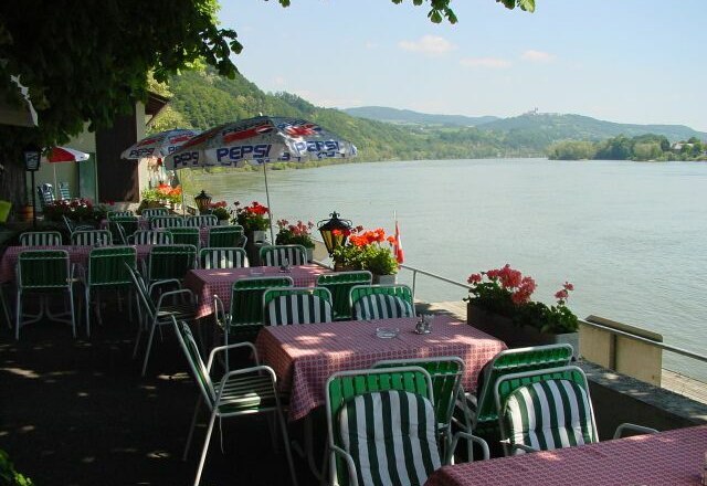 Terrasse eines Restaurants mit Blick auf die Donau, Tische mit rot-weiß karierten Tischdecken, grüne Stühle und Pepsi-Sonnenschirme.