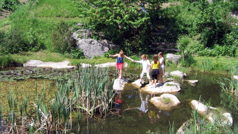 Kinder spielen an einem Teich im Gr&uuml;nen mit Seerosen und Schilf.
