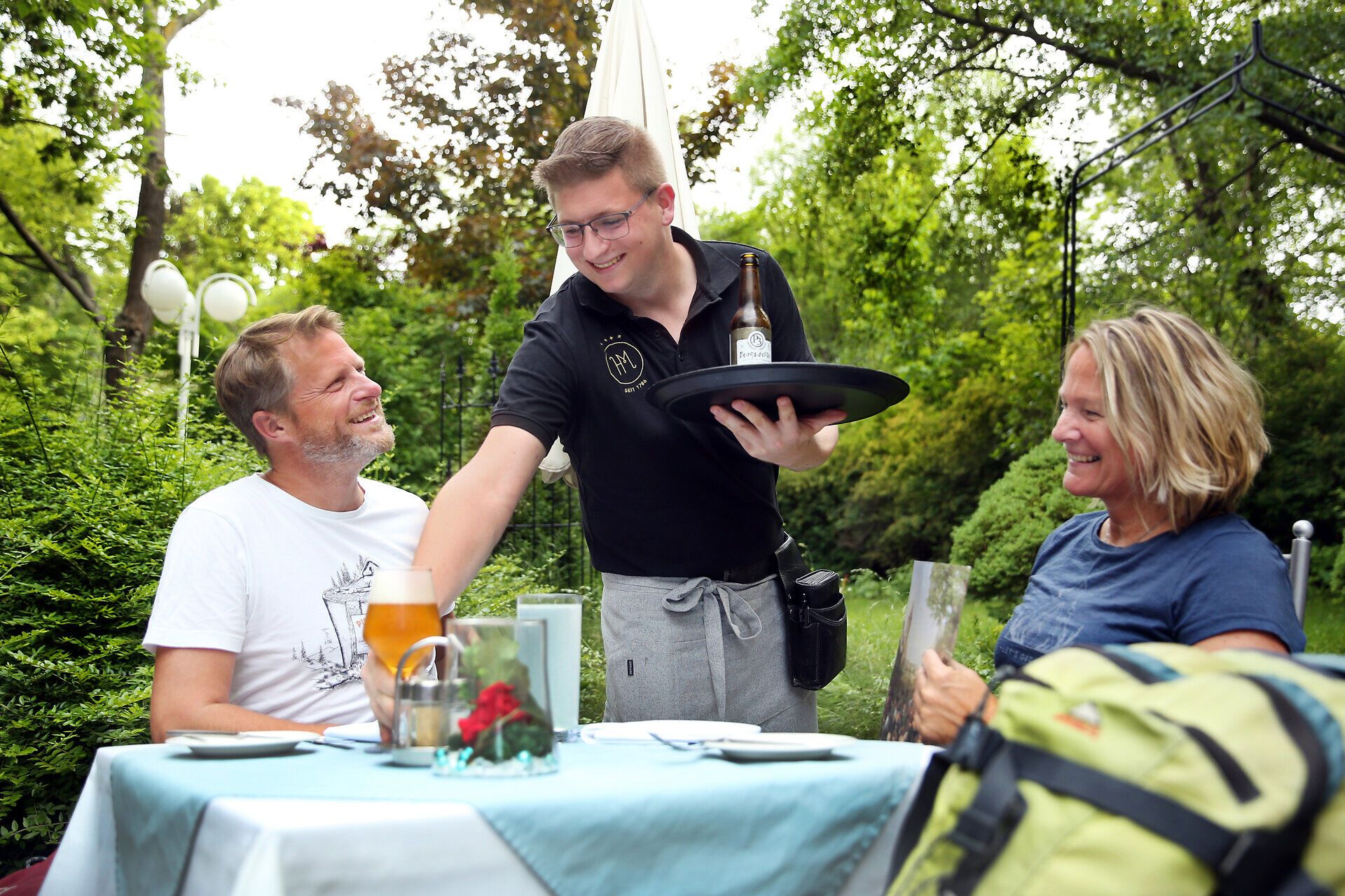 Inmitten einer idyllischen Gartenlandschaft genießen zwei Gäste ein köstliches Essen, während ein aufmerksamer Kellner ihnen eine Flasche Wein serviert. Die sanfte Brise und das Zwitschern der Vögel schaffen eine entspannte Atmosphäre, die zum Verweilen einlädt.