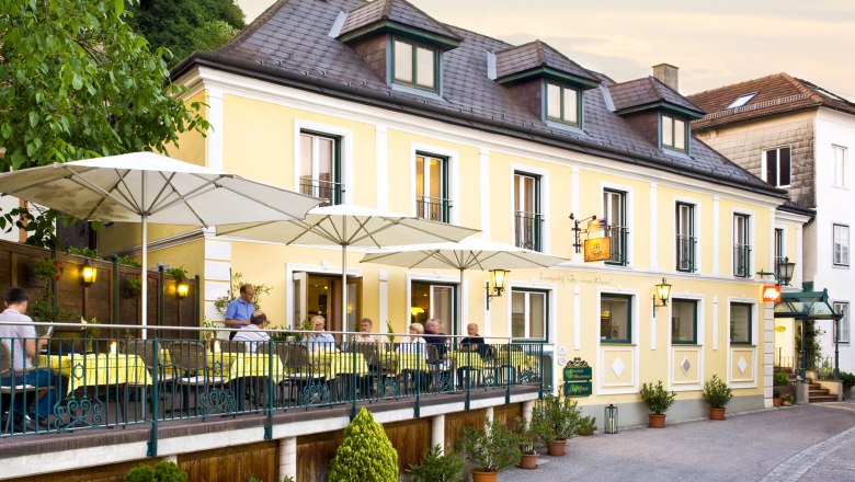 Ein gelber Landgasthof mit Terrasse und Sonnenschirmen, auf der Menschen sitzen.