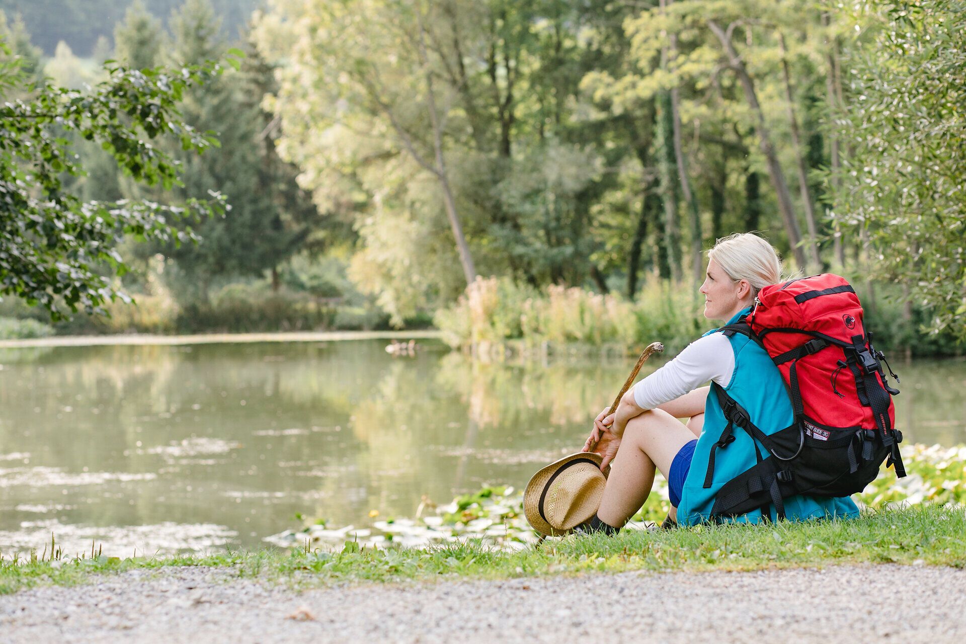 Eine Frau genießt eine wohlverdiente Pause am Ufer eines ruhigen Gewässers, umgeben von der üppigen Natur des Mostviertels. Ihr Rucksack zeugt von einem abenteuerlichen Tag voller Erkundungen und Pilgerreisen. Die sanfte Brise und das Plätschern des Wassers schaffen eine friedliche Atmosphäre, die zum Verweilen einlädt.