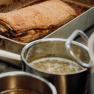 Knuspriger Schweinsbraten in einer Metallpfanne, daneben Töpfe mit Soße.