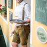 Ein Mann in traditioneller Kleidung lehnt an einer Wand und h&auml;lt ein Tablett mit einem Glas Bier.