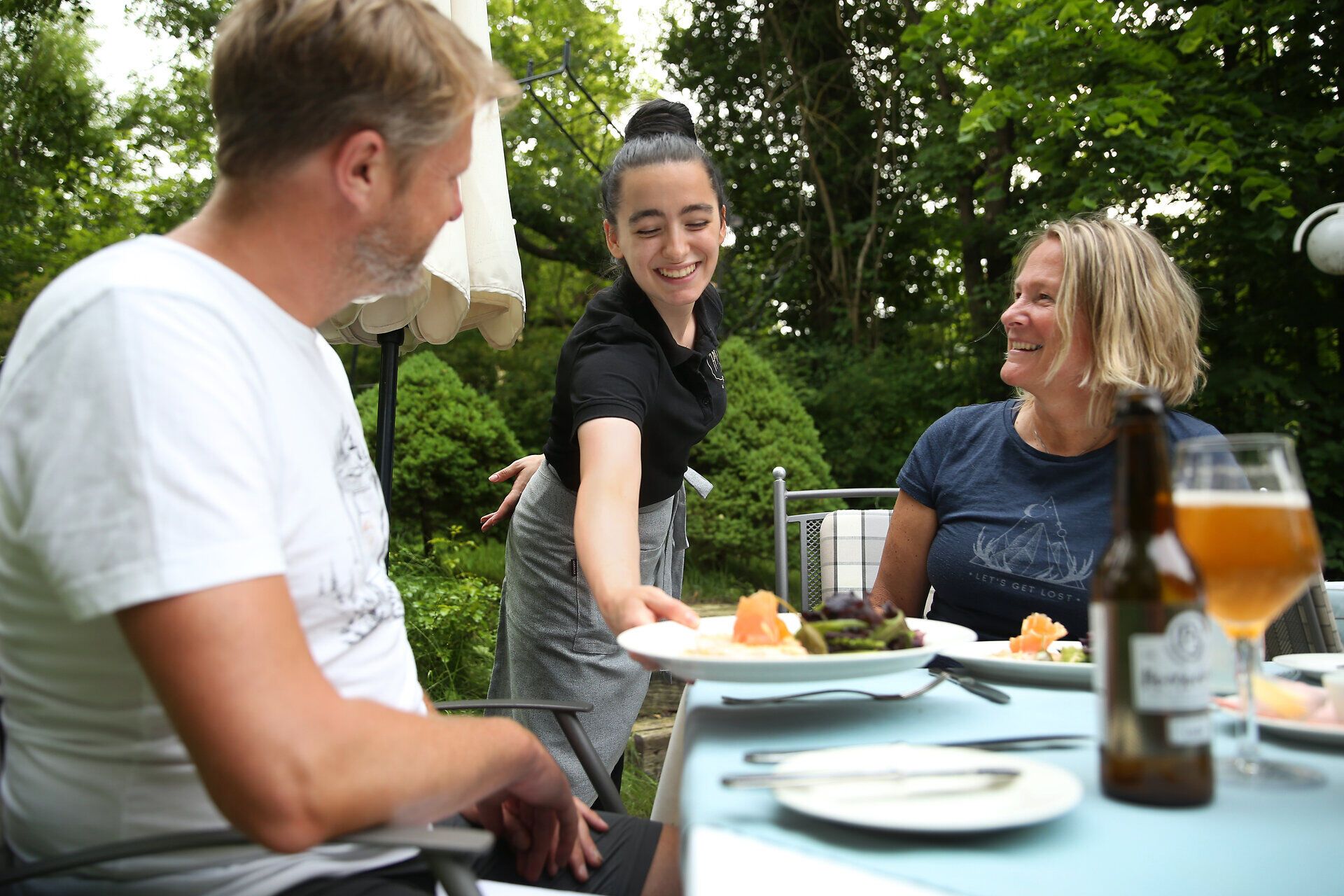 Inmitten der grünen Natur genießen Gäste eine köstliche Mahlzeit, während die Sonne sanft durch die Bäume scheint. Die freundliche Bedienung bringt mit einem Lächeln die Speisen, die die Vorfreude auf einen entspannten Tag in der Natur wecken. Hier wird die Verbindung zur Umgebung spürbar, während man die Ruhe und Schönheit der Landschaft auf sich wirken lässt.