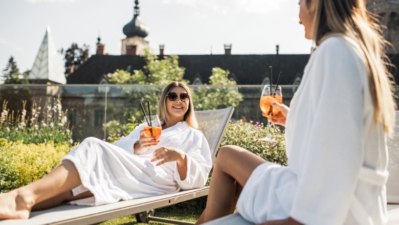 Zwei Frauen in Bademänteln entspannen auf Liegestühlen mit Getränken vor einem Schloss.