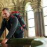 Ein Mann mit Rucksack beugt sich &uuml;ber ein Wasserbecken in einem historischen Geb&auml;ude mit bunten Glasfenstern.