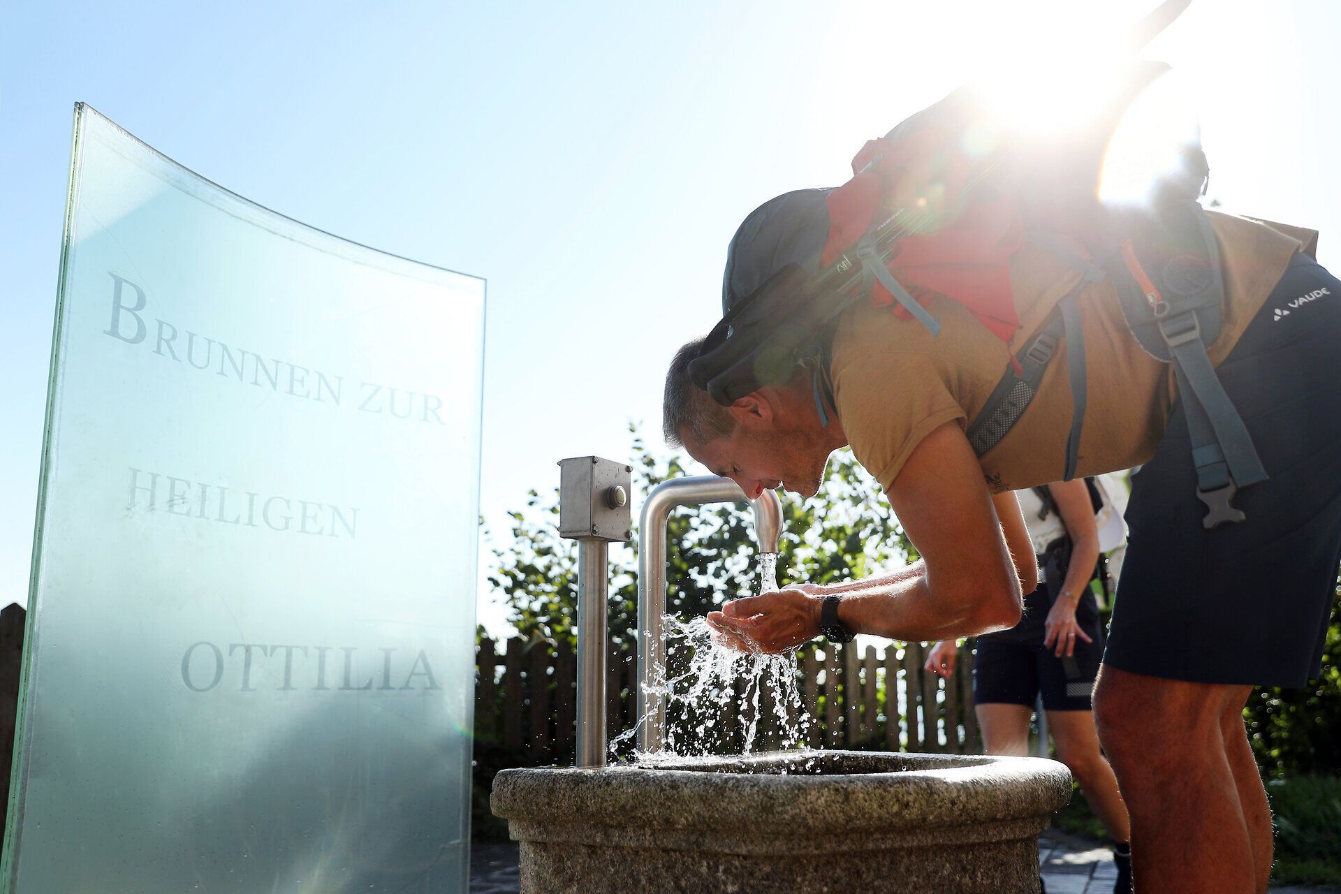 Ein Wanderer beugt sich über den sprudelnden Brunnen, um sich mit frischem Wasser zu erfrischen. Die strahlende Sonne hinter ihm taucht die Szene in ein warmes Licht und verstärkt das Gefühl von Freiheit und Abenteuer in der Natur. Umgeben von der malerischen Landschaft des Mostviertels wird jeder Schluck zu einem Genuss.