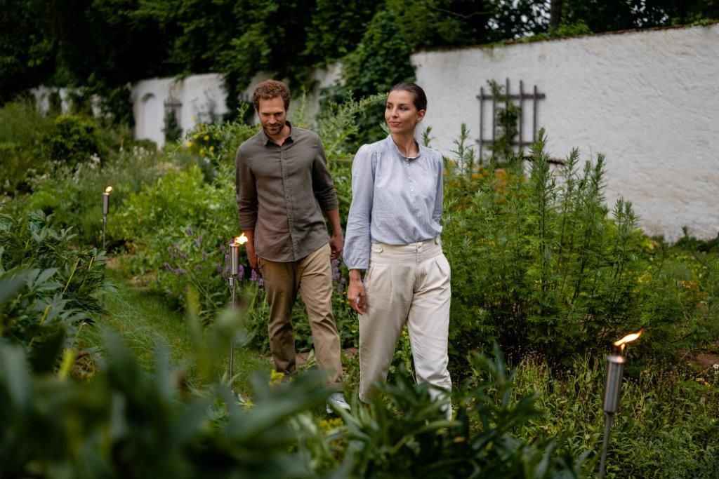 Ein Mann und eine Frau spazieren durch einen grünen Garten, umgeben von Pflanzen.