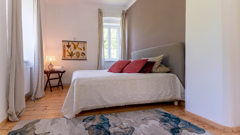 Schlafzimmer mit Bett, roten Kissen, Teppich und Fenster mit Vorhängen.