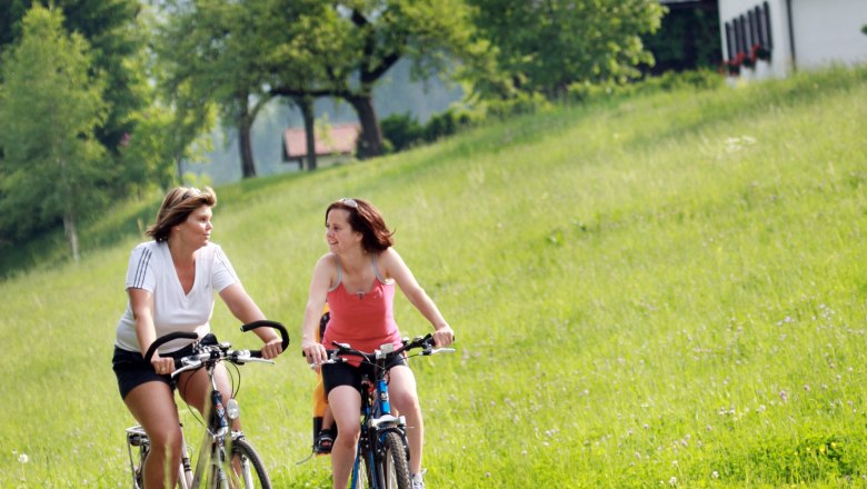 Zwei Frauen fahren auf Fahrrädern durch eine grüne Landschaft mit Hügeln und Bäumen im Hintergrund.