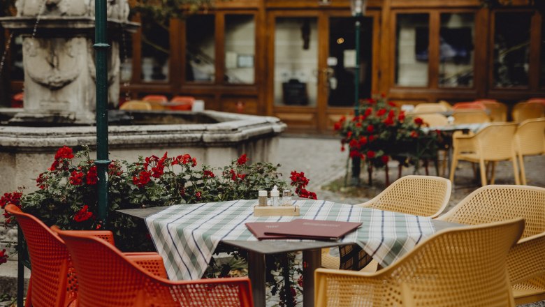 Ein Gastgarten mit bunten Stühlen, einem Tisch mit Tischdecke und einem Brunnen im Hintergrund.