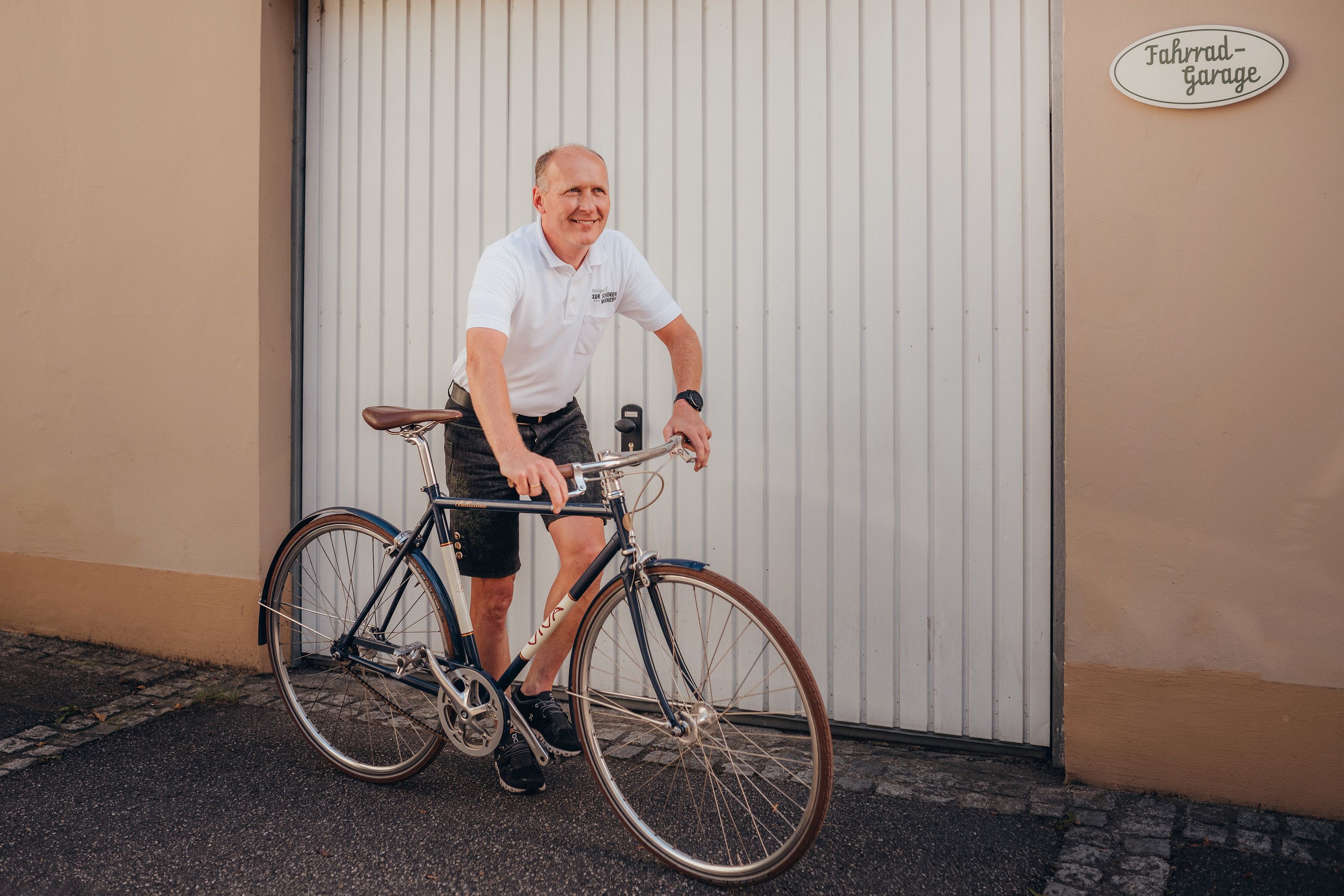 Mann in weißem Polohemd fährt Fahrrad vor Garagentor.