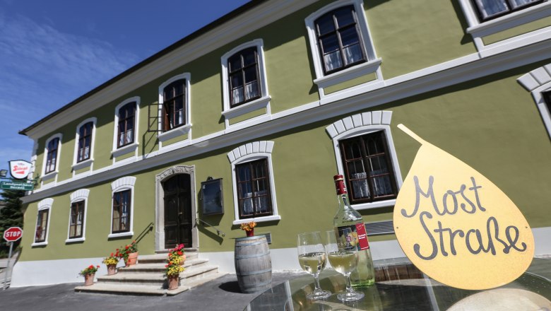 Gr&uuml;nes Geb&auml;ude mit wei&szlig;en Fenstern, davor ein Tisch mit Weinflasche und Gl&auml;sern, Schild mit Aufschrift 'Most Stra&szlig;e'.
