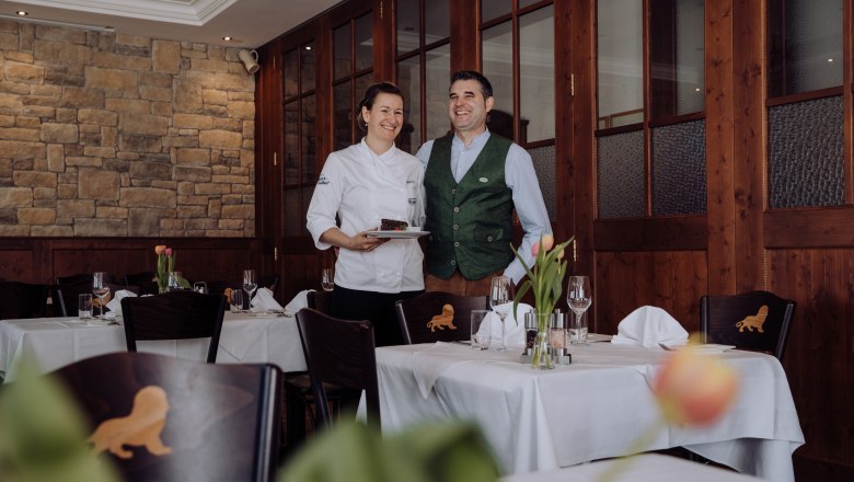 Eine Chefköchin und ein Mann in traditioneller Kleidung stehen lächelnd in einem eleganten Restaurant.