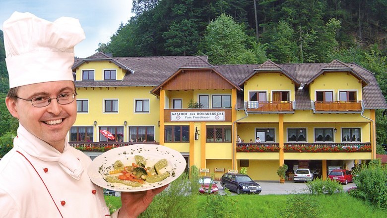 Ein Koch präsentiert ein Gericht vor einem gelben Gasthof namens 'Zur Donaubrücke'.