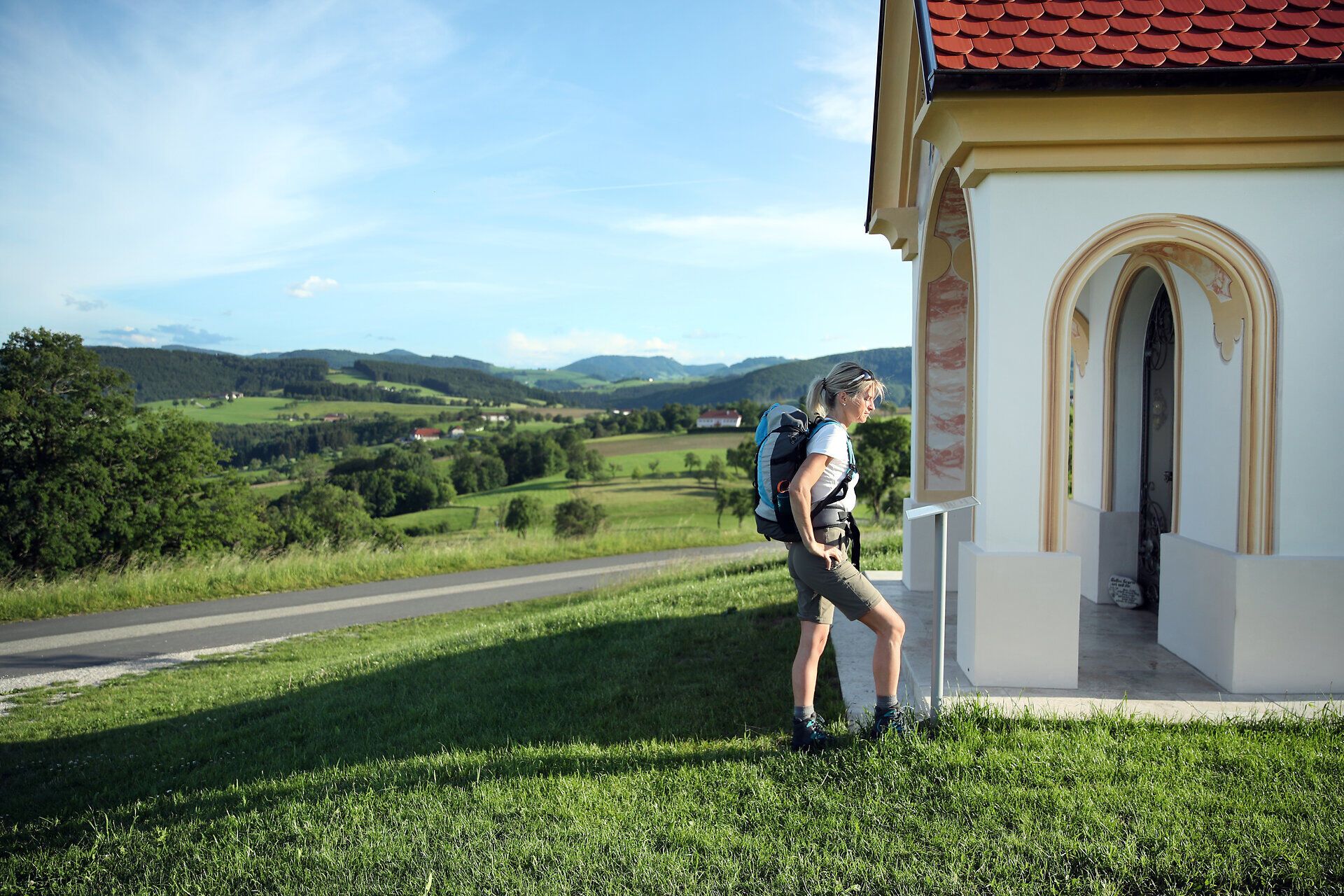 Pilgerin steht bei kleiner Kapelle entlang des Pilgerwegs.