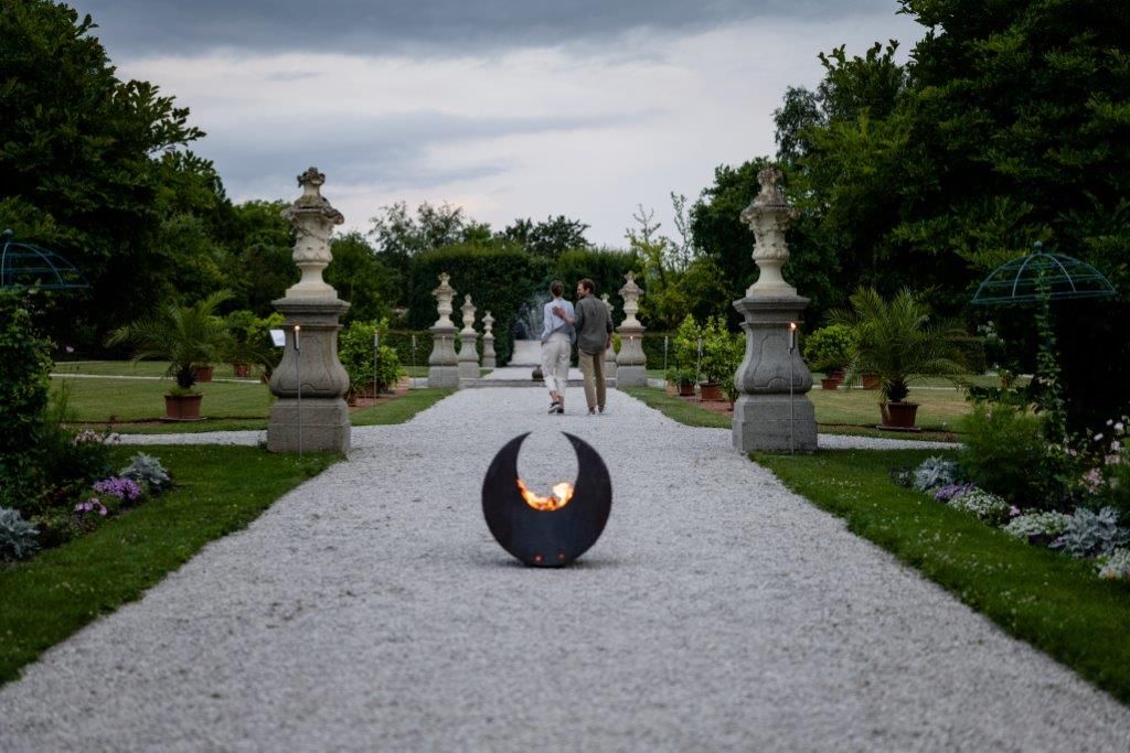 Ein Kiesweg im historischen Hofgarten des Stifts Seitenstetten mit Skulpturen und Pflanzen, im Vordergrund eine Feuerstelle.