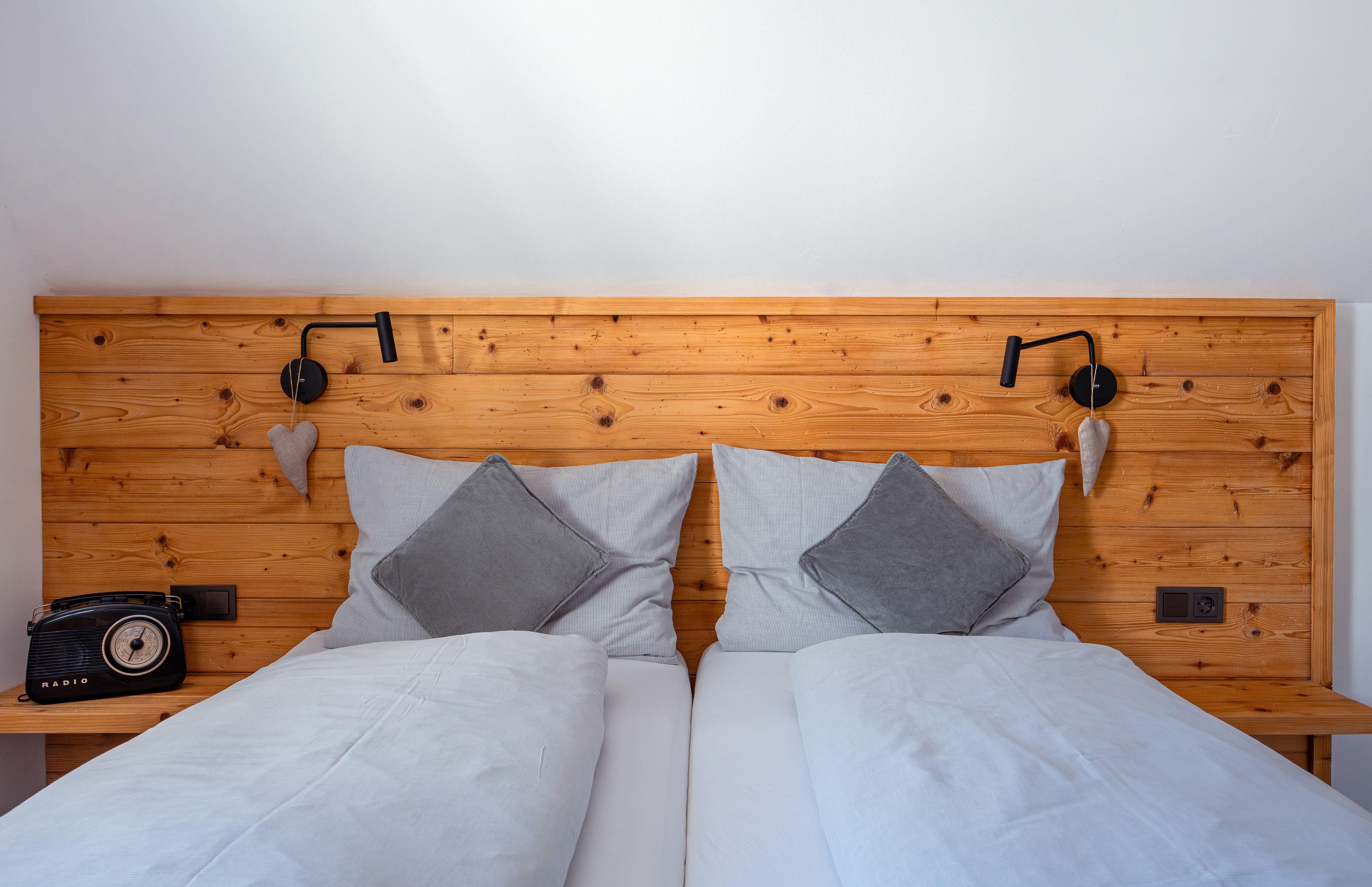 Gemütliches Doppelbett mit Holzrückwand, grauen Kissen und Decken. Links ein Retro-Radio, rechts eine Leselampe und Herzdeko.