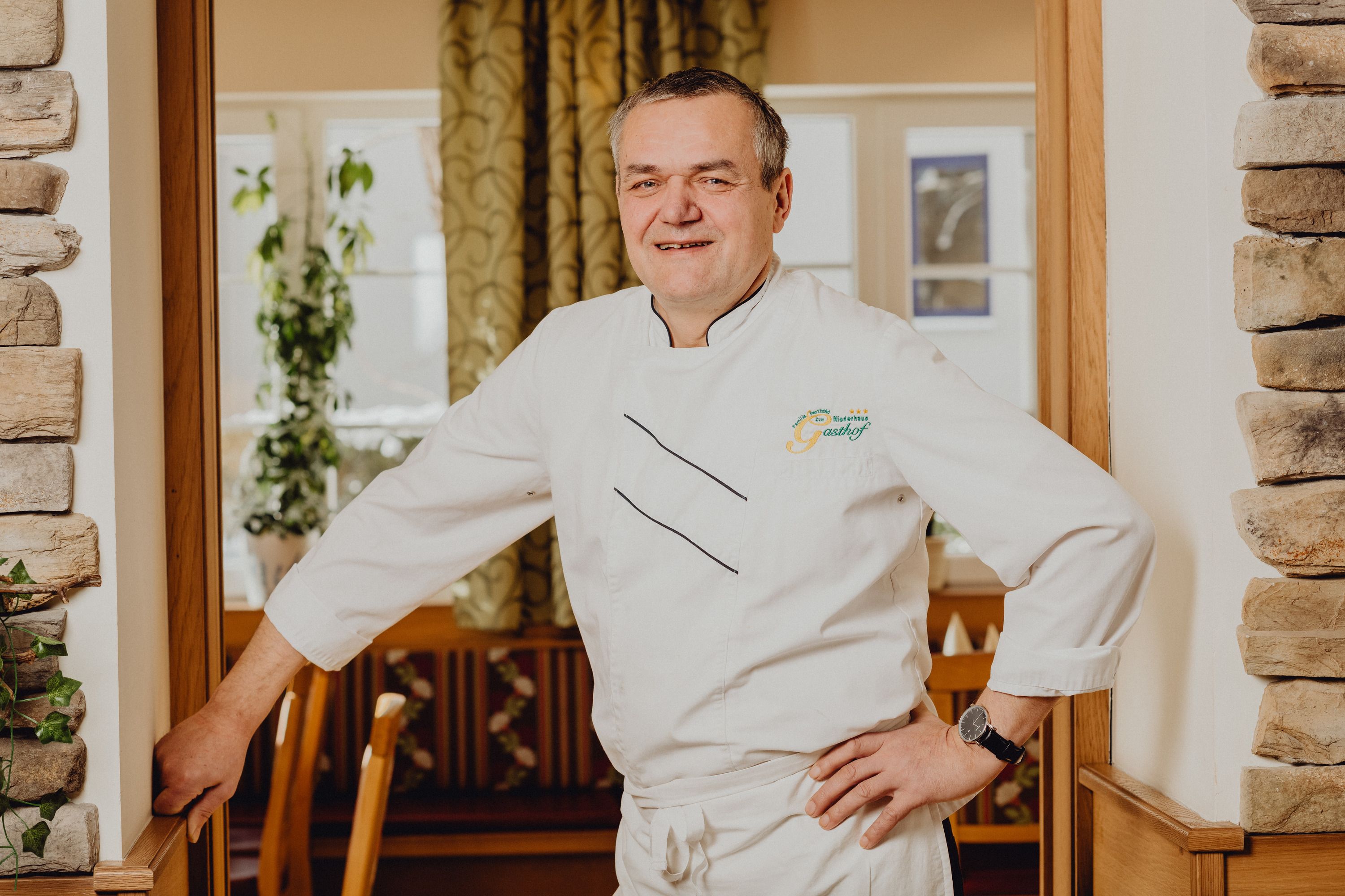 Ein Mann in weißer Kochuniform steht lächelnd in einem Restaurant.