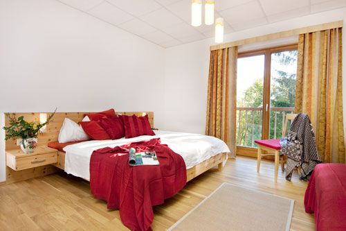 Helles Schlafzimmer mit Doppelbett, roten Kissen und Decke, Holzboden und großem Fenster mit Vorhängen.