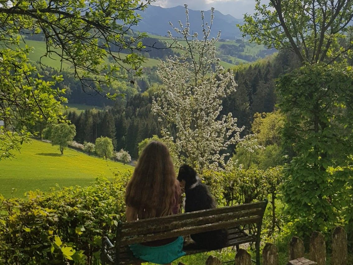 Eine Frau sitzt mit einem Hund auf einer Bank und blickt auf eine grüne Landschaft mit blühenden Bäumen und Bergen im Hintergrund.