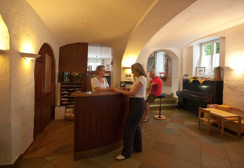 Eine Hotelrezeption mit zwei Frauen im Gespräch und einem Mann, der auf einem Hocker sitzt.