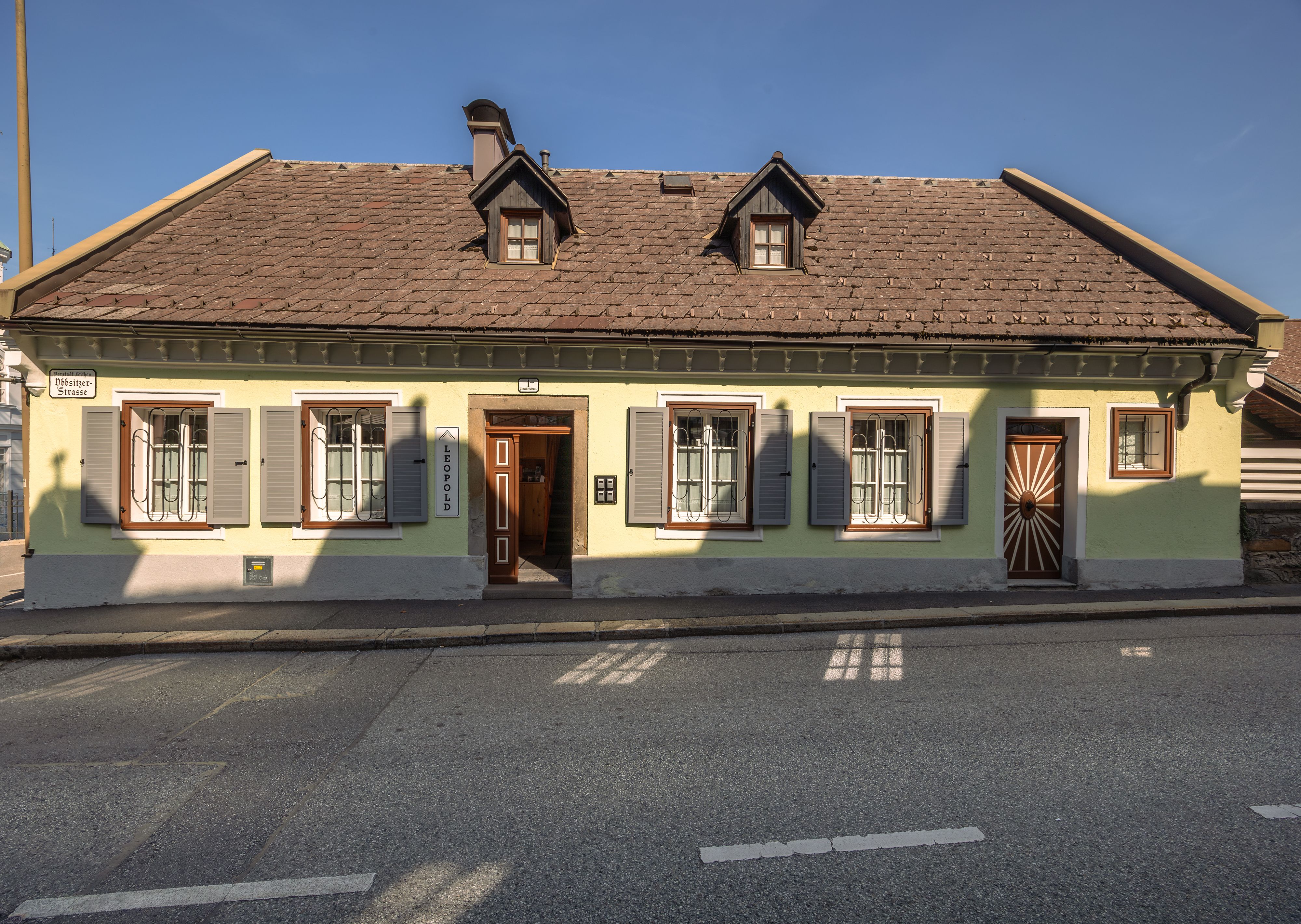 Ein traditionelles, gelbes Haus mit braunem Dach und grünen Fensterläden an einer Straße.