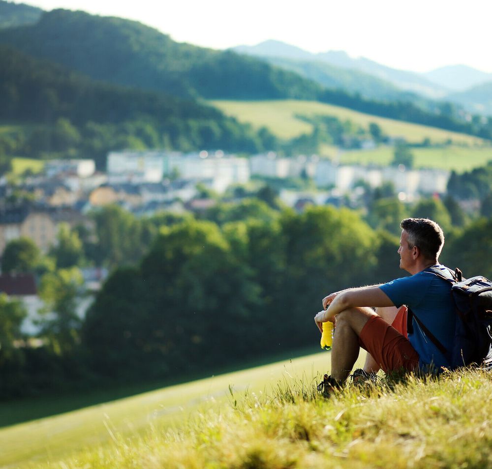 Ein Wanderer genießt die atemberaubende Aussicht auf die sanften Hügel und das malerische Dorf, während die warmen Sonnenstrahlen den Sommer in den Bergen erhellen. Die frische Luft und die ruhige Umgebung laden dazu ein, die Seele baumeln zu lassen und die Schönheit der Natur zu erleben.