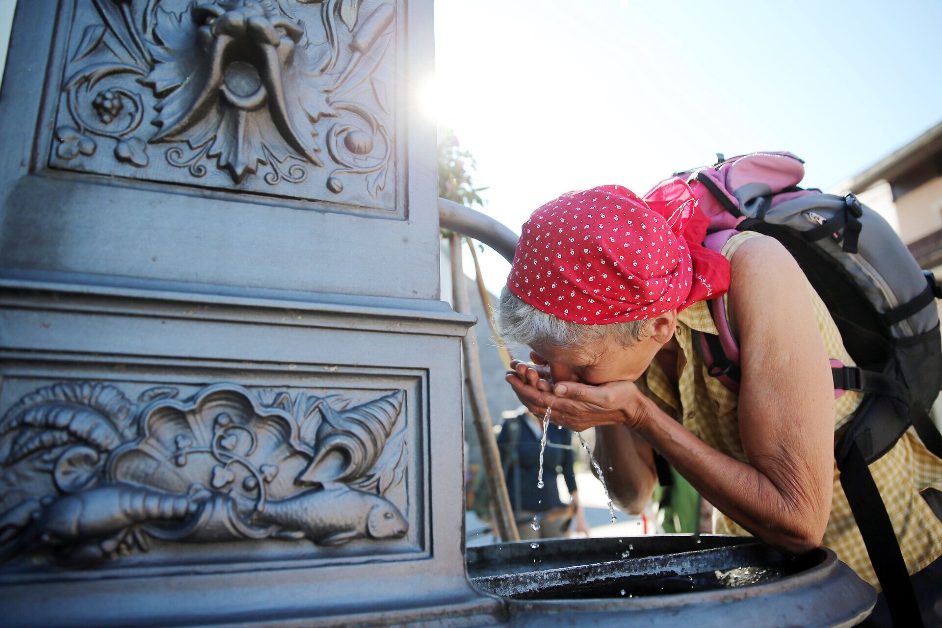 Eine Frau genießt eine erfrischende Trinkpause an einem historischen Brunnen, während die warmen Sonnenstrahlen sanft auf sie herabfallen. Umgeben von der malerischen Landschaft des Mostviertels, spürt sie die Ruhe und den Frieden, die das Pilgern auf der Via Sacra mit sich bringt.