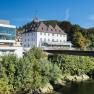 Ein modernes Geb&auml;ude neben einem historischen Schloss, umgeben von B&auml;umen und einem Fluss, unter blauem Himmel.