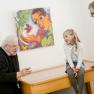 Zwei Pater sprechen mit zwei Kindern in einer Galerie vor einem bunten Gemälde.