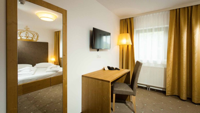 Ein Hotelzimmer mit einem Doppelbett, einem Schreibtisch mit Stuhl, einem Fernseher an der Wand und gelben Vorh&auml;ngen.