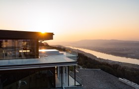 Modernes Gebäude mit Glasfassade bei Sonnenuntergang, Blick auf Fluss und Landschaft.