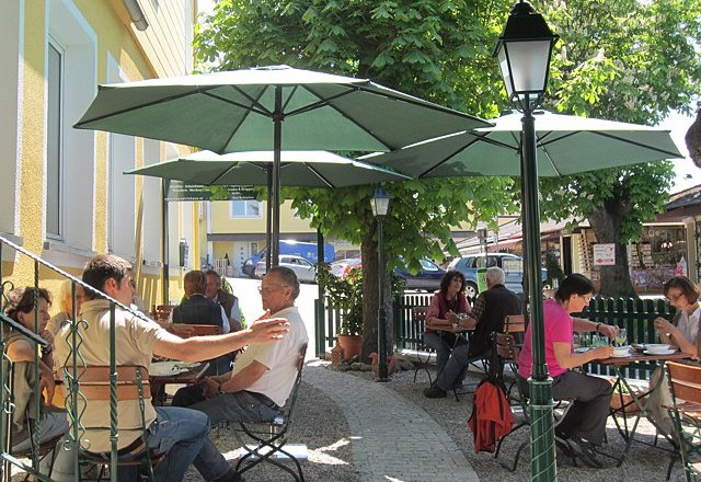 Menschen sitzen in einem sonnigen Gastgarten mit grünen Sonnenschirmen.