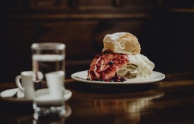Ein Teller mit einer Klostercremeschnitte, garniert mit roten Beeren, steht auf einem Holztisch. Im Hintergrund sind ein Glas Wasser und zwei Espressotassen zu sehen.