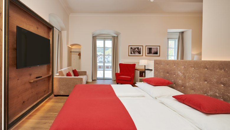 Hotelzimmer mit Doppelbett, roter Bettwäsche, Sofa, rotem Sessel und großem Fenster.