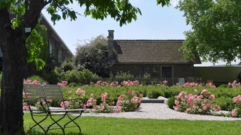 Ein altes Gebäude in einem Garten mit blühenden Rosen und einer Bank im Vordergrund.
