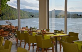 Innenansicht eines Restaurants mit Blick auf einen Fluss und grüne Hügel.