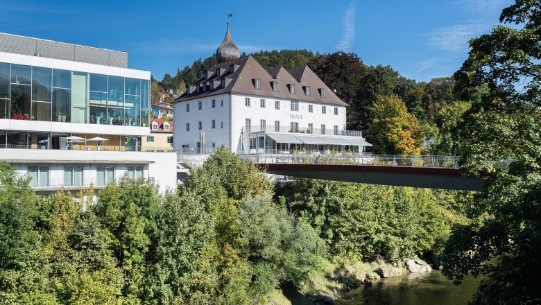 Schloss an der Eisenstraße, © Stixenberger Ein modernes Gebäude neben einem historischen Schloss, umgeben von Bäumen und einem Fluss, unter blauem Himmel.