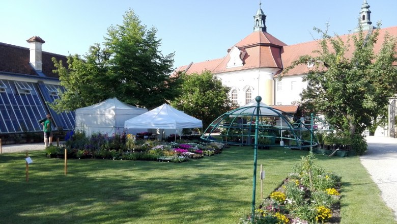 Garten mit Blumenbeeten, weißen Zelten und historischem Gebäude im Hintergrund.